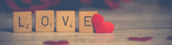 'LOVE' is spelt out with scrabble letters standing up, with a red heart at the end of the 'e'. Red hearts are also flat on the table which are out of focus