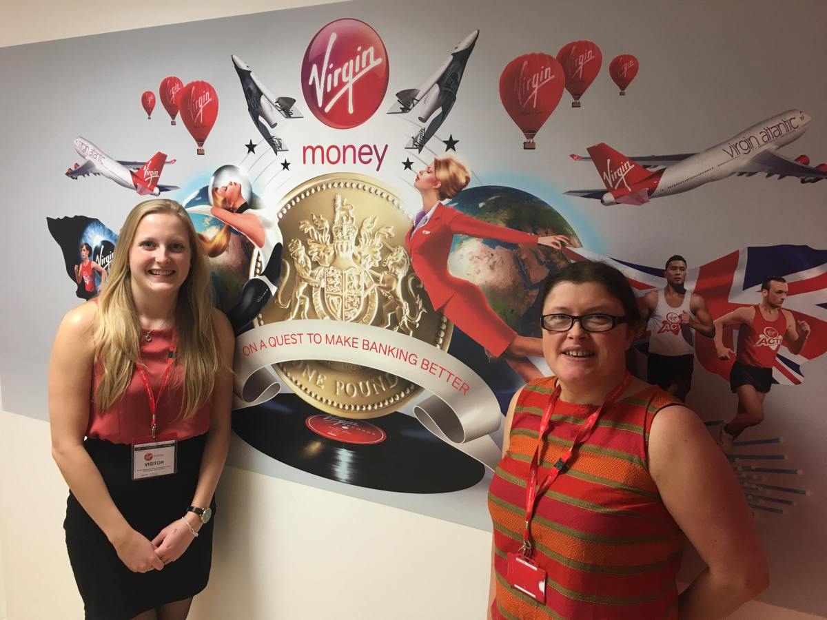 DB standing to left, and Vick from Virgin Money Bank standing to the right. Between them on the wall is a big mural photo collage with lots of Virgin services and products with their motto 'On a quest to make banking better'