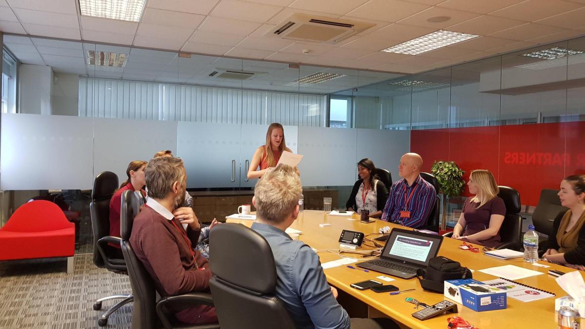 Deafie Blogger standing at the far end of a long oval conference table, holding presentation slides and giving a talk to members of staff from Virgin Money Bank who are sitting around the table. There is also glass walls around the room and a laptop on the table.