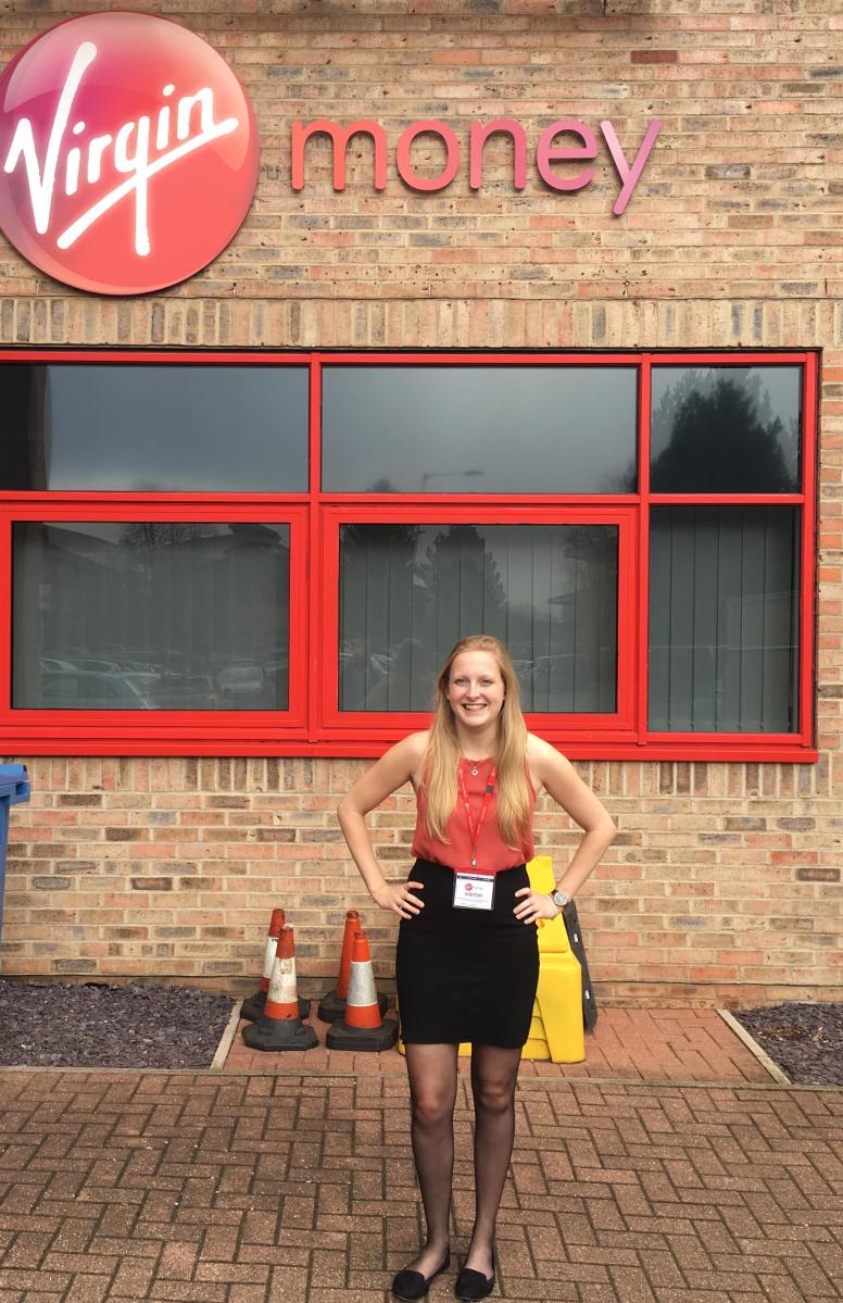 Deafie Blogger standing outside the building of Virgin Money Bank. Virgin Money Logo is above a red window which DB is standing in front of. Wearing a coral sleeveless top and black skirt. The building wall is brick.