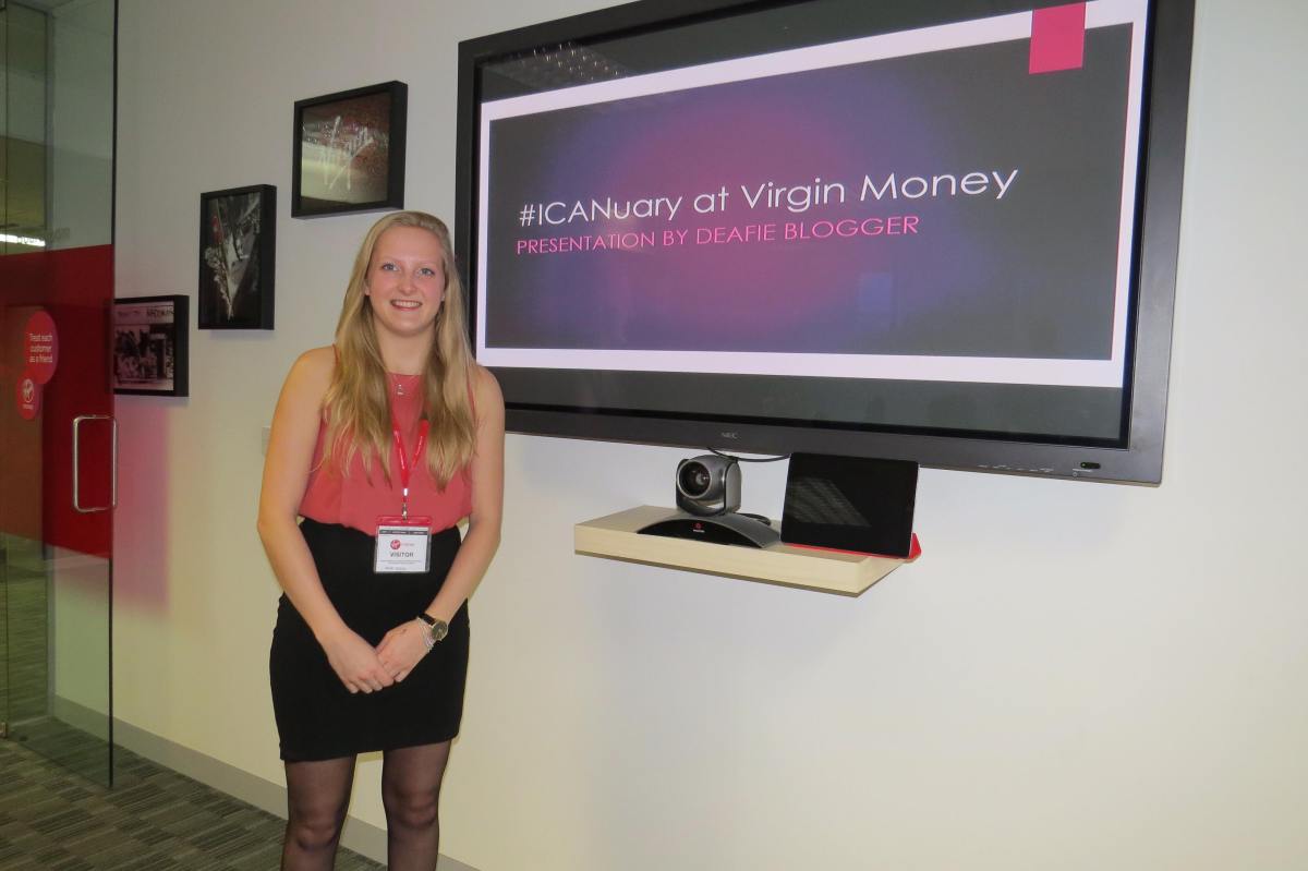 DB standing to the left of a TV screen with her presentation. Main slide is purple and says #ICANuary at Virgin Money, Presentation by Deafie Blogger