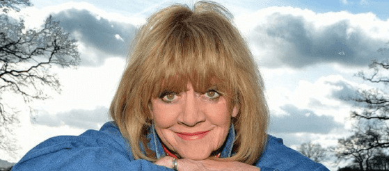 Close up head and shoulders shot of Amanda Barrie wearing a blue denim shirt smiling directly into the camera with a blue and grey sky behind her`