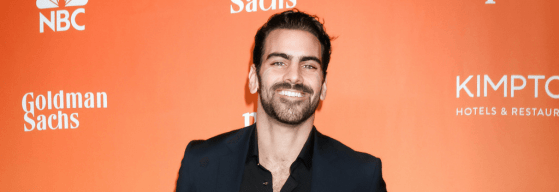 A photo of deaf model Nyle Di Marco smiling, standing against an orange background for an event, wearing a black suit and shirt.