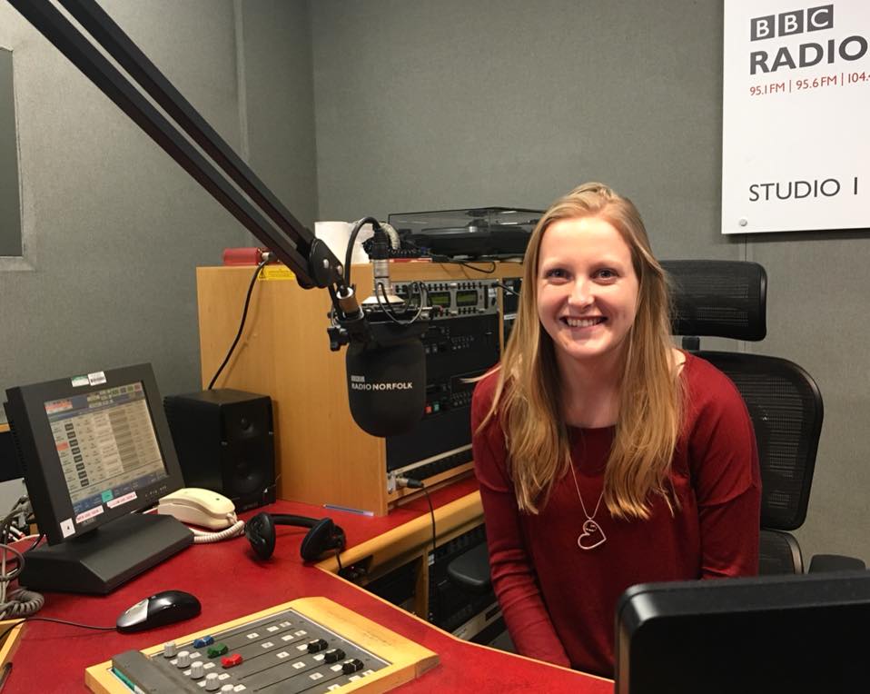 Deafie Blogger smiling sitting in a Radio Recording Studio wearing a red top, heart necklace with recording panels and microphones in front of her