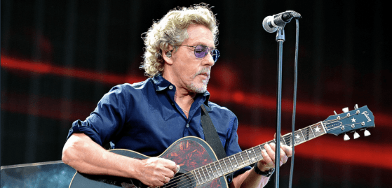 The Who's Roger Daltrey playing guitar on stage with a microphone in front of him. Also wearing protective earpieces