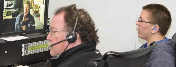 Johanna on the right, wearing black hearing aid with blue earmould and glasses, with a blue hoody is facing left staring at a computer which has an interpreter on. On the left is a man wearing a headset and microphone, with glasses, also looking in the same direction at a computer