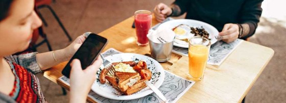 Couple at a small table in a restaurant, camera angle looking down at the table which the girl is holding her phone and there are two plates, both with a fry up on, and one has orange juice, the other has cranberry or tomato juice.