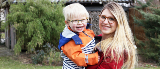 Photo of Mother (right) holding her son in her arms (left). Mother has long blonde hair and is wearing curved glasses and a red coat. Son is wearing a blue and white striped coat with orange shoulders and arms and a blue hood, also has short blond hair and wearing rectangular glasses. Both are smiling and standing in a garden.