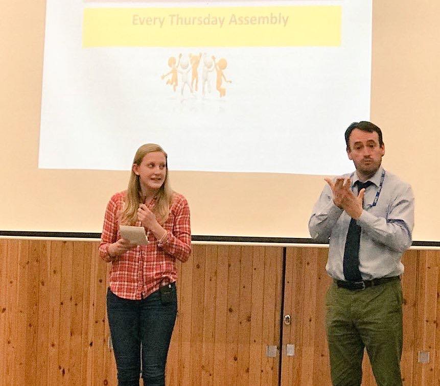 Deafie Blogger giving her speech holding a notepad left, wearing a red chequered top and blue jeans with the Interpreter on the right signing