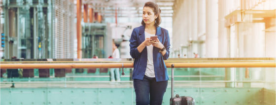 Picture of a woman on her phone but looking to her left with a suitcase by her right side with the handle pulled up. The lady is wearing a blue blazer, blue jeans and white top.