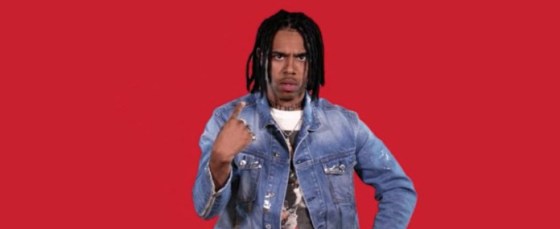 Photo of a man standing in front of a bright red background with black hair, denim jacket and a black and white printed top pointing at himself with an 'unsure' look on his face