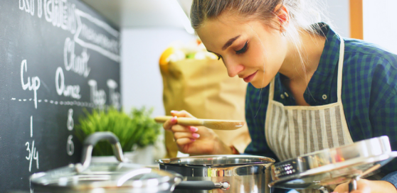Woman smelling a saucepan on the hob in the kitchen. To her left is another saucepan and also a chalkboard with written measurements on it and also in the background is a green herb plant and next to it is a brown carrier bag but they are blurred