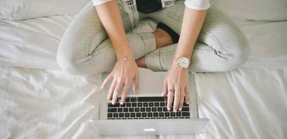 Birds eye view of girl typing on a laptop while sitting on a bed with legs crossed