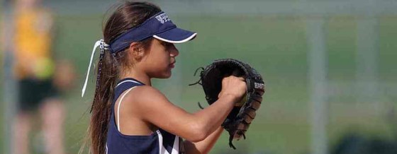 Photo of a young girl with a baseball mitt facing sideways looking straight in that direction. She is wearing a blue and white athletic top and blue and white cap with her hair tied back