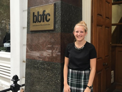Image of myself standing outside the UK Cinema Association office. I am wearing a black short sleeved top with embellished neckline, and a green, black and white chequered skirt. I am smiling and my hair is in a bun.