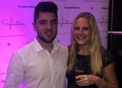 A couple dressed up smart smiling with Signature awards backdrop, holding a prosecco glass