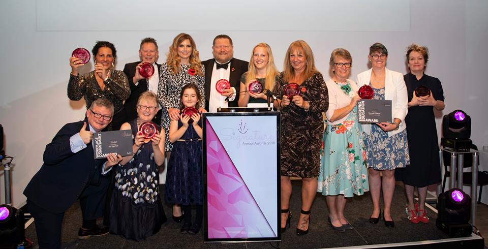 10 Award Winners on the stage smiling with their trophies along with Pip Tomson, I am in the middle next to Craig Crowley MBE in front of a podium.