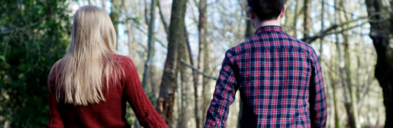 Couple holding hands in the woods, photo taken from behind, girl on left long blonde hair wearing red jumper, boy on right short black hair wearing blue and red chequered shirt