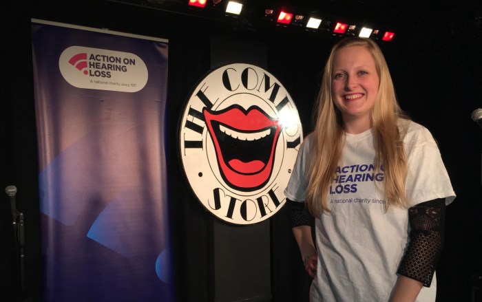 Me smiling wearing white Tshirt with AOHL logo on. In background is comedy store logo and also AOHL banner
