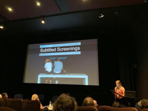 Big screen photo of two girls watching subtitles on a screen. Text says 'Subtitled Screenings'. I can standing at the bottom right giving my presentation.