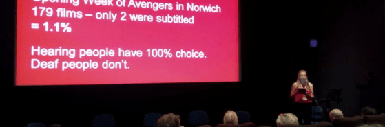 My #SubtitledCinema presentation at the Independent Cinema Office - I am holding a microphone speaking to a crowd, behind me is my powerpoint with stats on