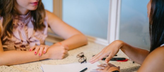 Two ladies chatting, sitting opposite, one has hand lifted up slightly. Glasses, pen and paper on the table